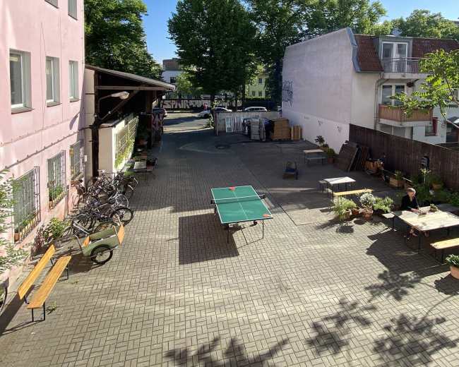 The image shows a sunny courtyard with bicycles parked, a ping pong table, and a person sitting at a table surrounded by potted plants.