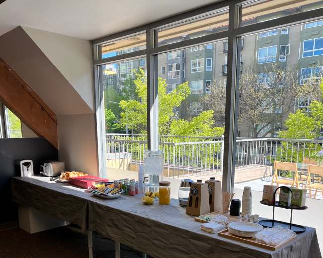 A long table is set up with food and drink items near large windows displaying greenery outside.