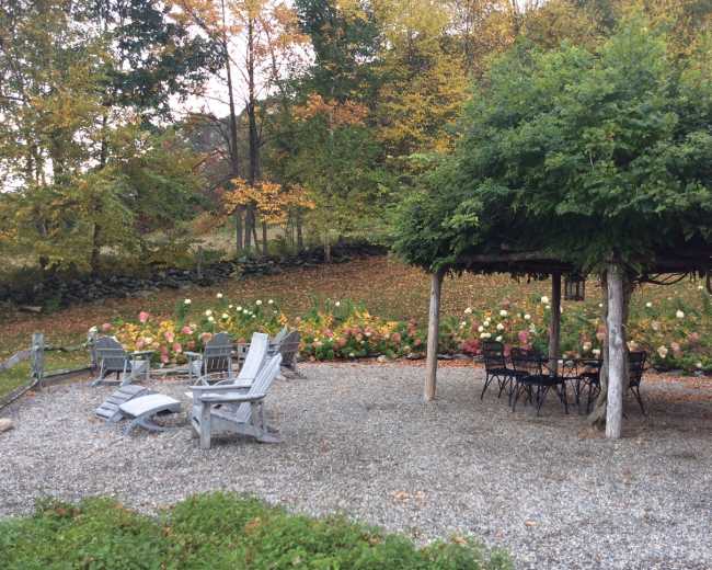A gravel patio area with wooden chairs and a table is surrounded by blooming flowers and trees in autumn.