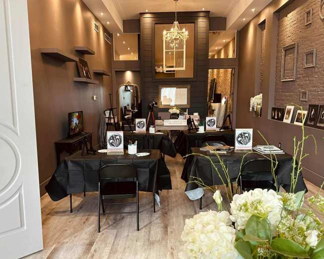 The image shows an elegantly arranged interior space featuring several tables with black tablecloths, decorative items on the walls, and a chandelier overhead.