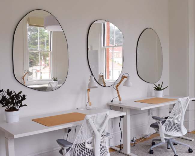The image shows a bright workspace featuring two white desks with ergonomic chairs, three oval mirrors on the wall, and potted plants, illuminated by natural light from a window.