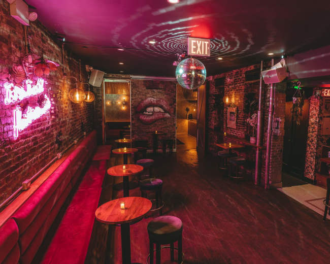 The image shows a dimly lit bar with exposed brick walls, circular tables, and a disco ball hanging from the ceiling.