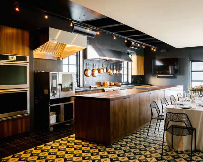 The image shows a modern kitchen with a large island, stainless steel appliances, and a dining table set for a meal.