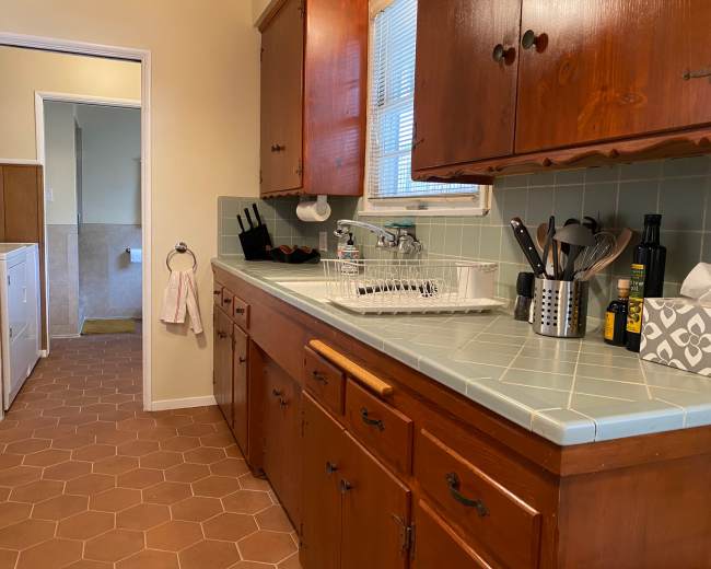 The image shows a kitchen with wooden cabinets, a light blue countertop, a sink with dishware, and a view into a hallway leading to a laundry room.