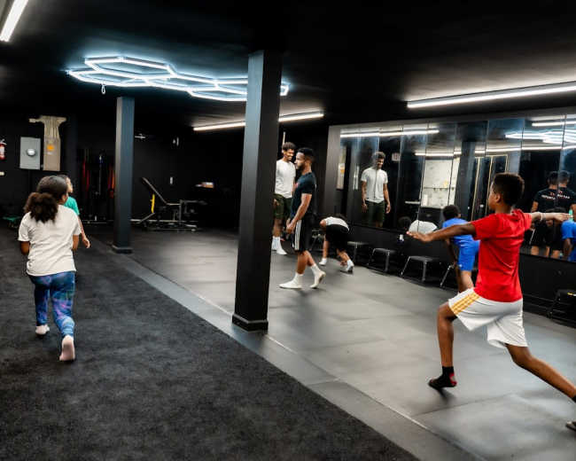 A group of children practice movement exercises in a dimly lit studio with black walls and a mirrored section.