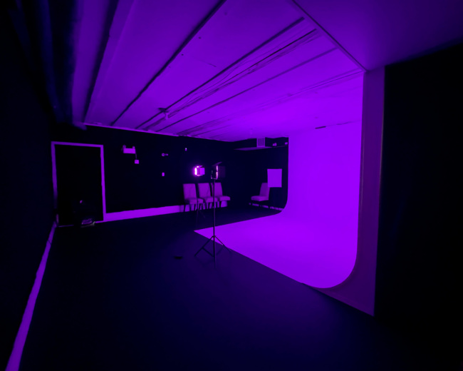 A studio space is illuminated in purple light, featuring a curved backdrop, a chair, and a tripod stand in the foreground.