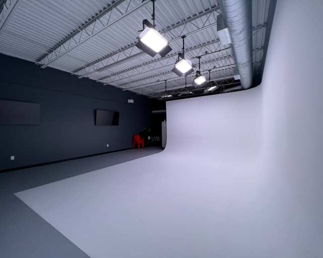 The image shows a spacious photography studio featuring a seamless white backdrop, several overhead lights, and minimalist decor.