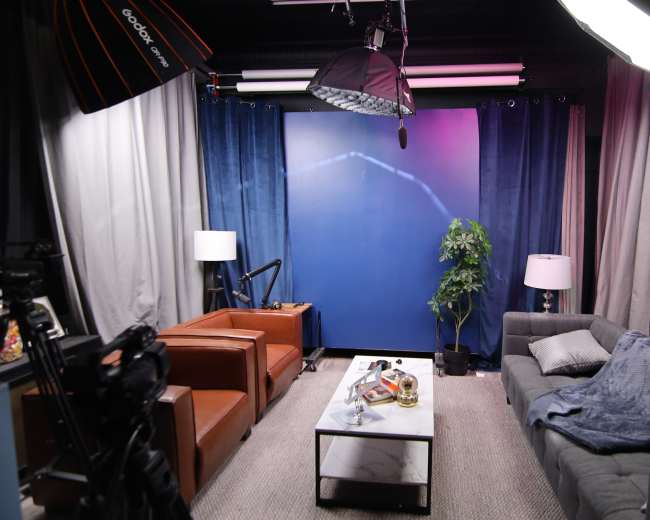 The image shows a well-lit studio setup with two armchairs, a sofa, a coffee table, and a backdrop of blue curtains and lighting equipment.