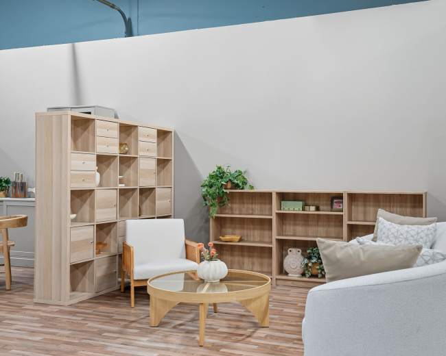 The image shows a modern interior space with a light-colored sofa and a round coffee table, surrounded by wooden shelves and a few potted plants.