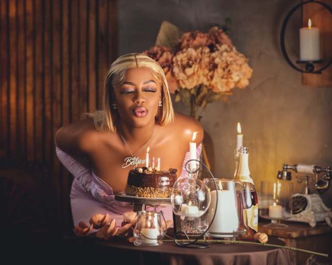 A woman in a pink dress leans over a chocolate cake adorned with candles on a table set with flowers and candles.