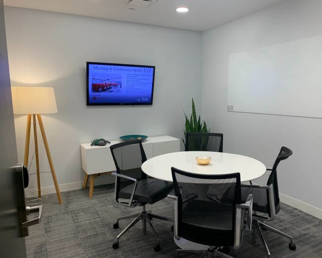The image shows a small meeting room featuring a round table with four chairs, a television mounted on the wall displaying information, a plant in the corner, and a floor lamp.