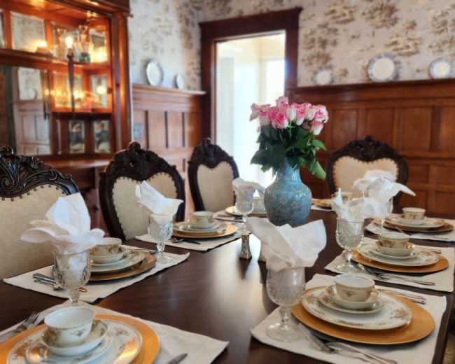 A dining table is elegantly set with fine china and crystal glassware, surrounded by ornate chairs, in a room featuring wooden paneling and floral wallpaper.