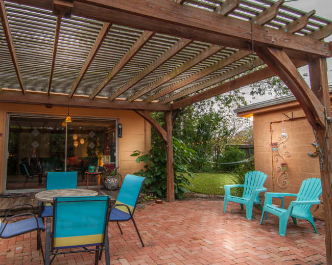 A covered patio features a circular table surrounded by six chairs and two lounge chairs, set against a backdrop of a well-maintained garden.