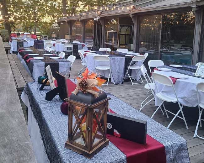 A series of tables are arranged outside, adorned with tablecloths and decorations, next to a building with large windows and hanging lights overhead.