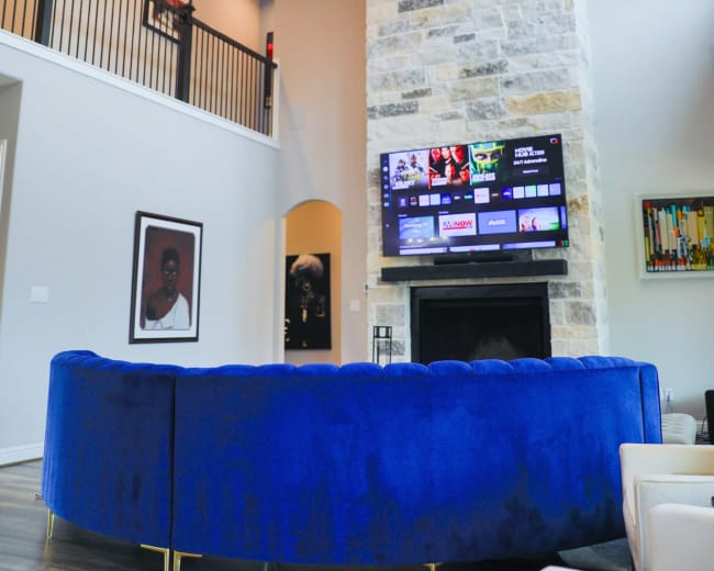 A modern living room features a curved blue sofa, a large stone fireplace, and a wall-mounted TV displaying various streaming options.