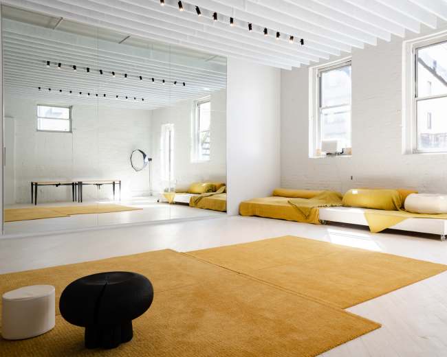 The image shows a minimalist interior space with a large mirror, a white sofa covered in yellow throws, and a beige rug on the floor.