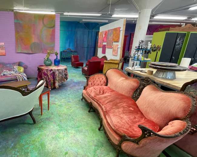 The image depicts a colorful interior space with eclectic furniture, including several vintage sofas and art-covered walls.