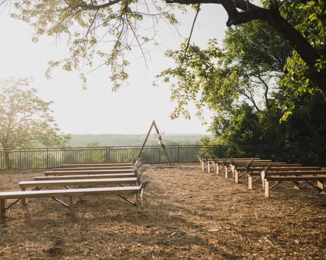 A rustic outdoor venue with wooden benches arranged in a semicircle facing an arched structure, surrounded by trees and overlooking a scenic view.