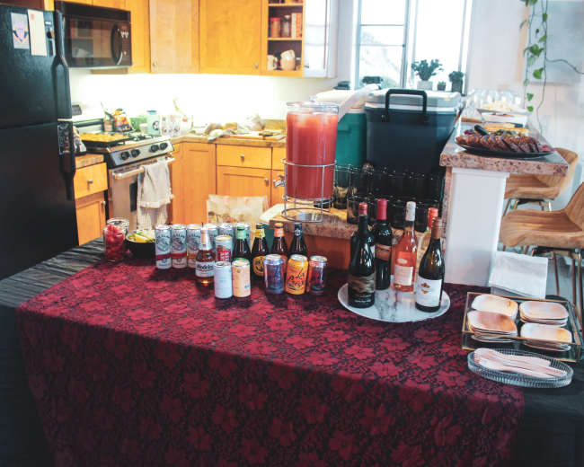 A table covered with a black tablecloth displays an array of drinks, including cans and bottles, in a kitchen setting.