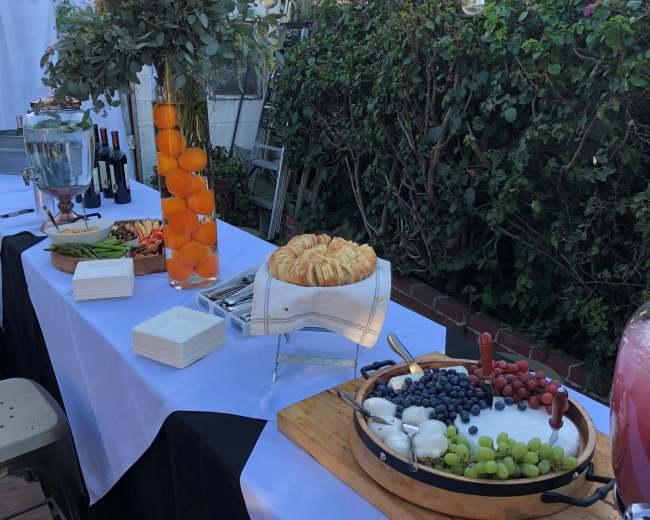 A long table is set outdoors with a variety of food items, including fruits, croissants, and cheeses, alongside drinks in glass containers.