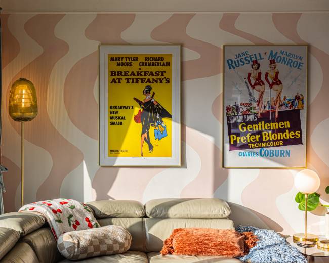 The image features a cozy living room with soft pink walls, two vintage movie posters framed on the wall, and a light-colored sofa adorned with decorative cushions.