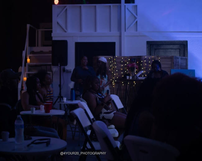 A group of people is seated at tables in a dimly lit venue, with some individuals performing on a stage in the background.