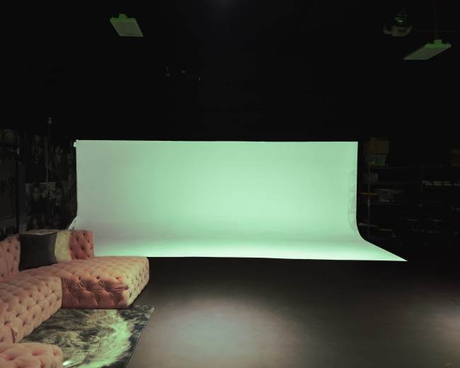 A studio space featuring a pink velvet sectional sofa, a patterned rug, and a large white backdrop.