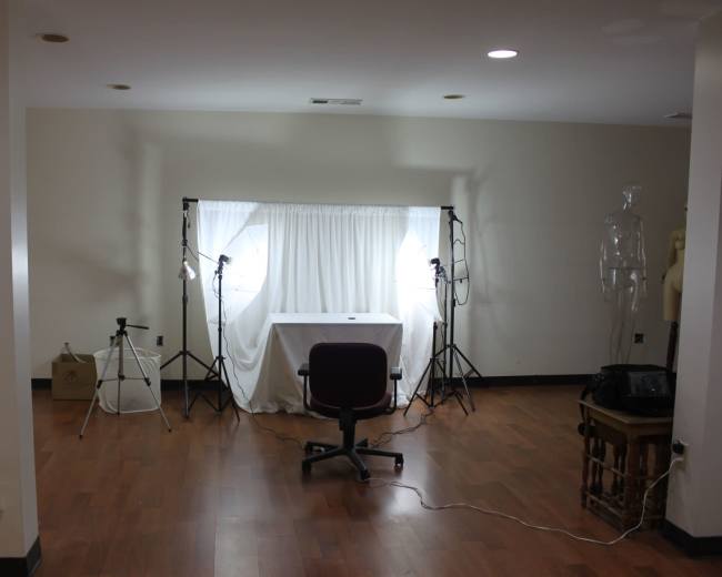 The image shows a spacious room with a table covered by a white cloth, flanked by two light stands and a chair, with a mannequin in the background.
