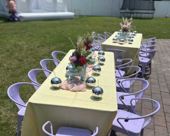 A long table set with decorative flowers and centerpieces is surrounded by purple chairs on a grassy area.