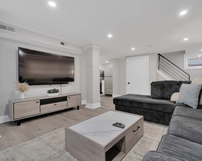 The photo shows a modern basement living room featuring a large sectional sofa, a flat-screen TV mounted on the wall, and a coffee table in front of a neutral-colored area rug.