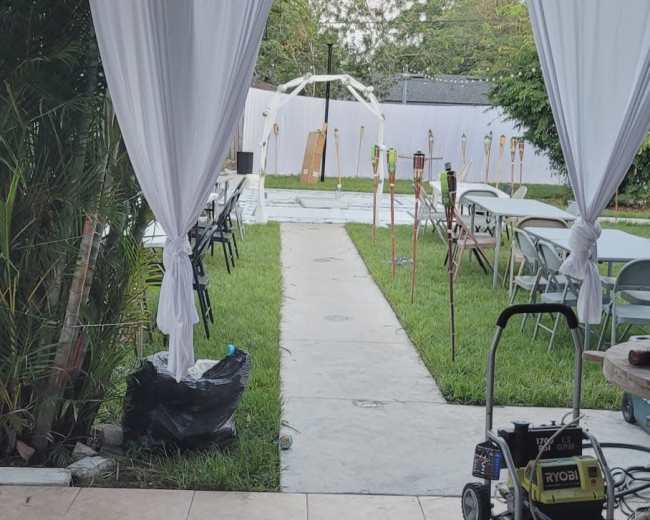 The image shows an outdoor space with a pathway lined by grass and tables set up for an event, viewed from a covered area with draped curtains.