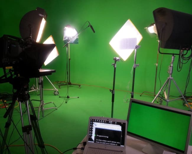 The image shows a studio setup with multiple lights on stands and a green screen as the backdrop, along with a laptop and monitor displaying a script.