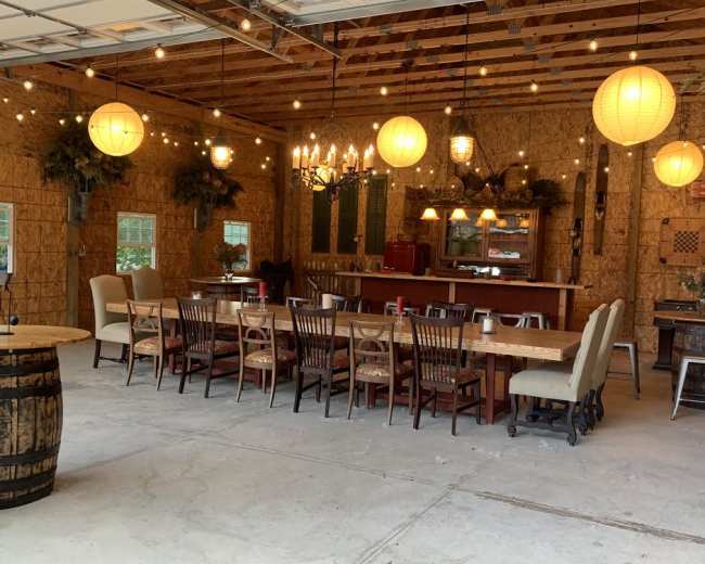 A spacious interior features a long dining table surrounded by chairs, with hanging lanterns and a chandelier illuminating the rustic decor.