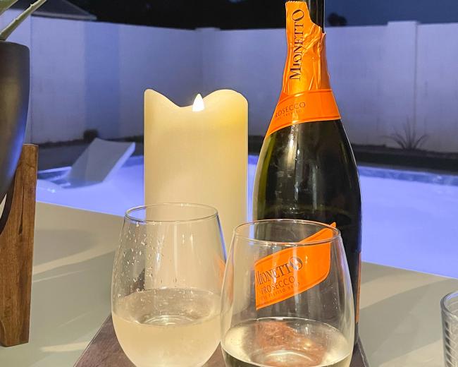 A bottle of Prosecco sits next to two half-filled glasses and a lit candle on a table, with a swimming pool and a night sky visible in the background.