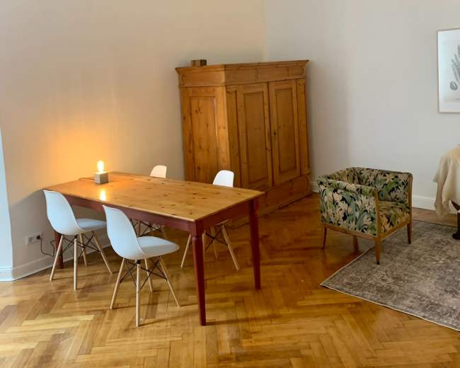 The image features a well-lit room with a wooden dining table and four chairs, a vintage cabinet, and two pendant lights hanging from the ceiling.