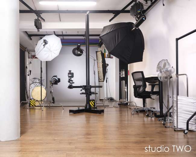 The image shows a photography studio equipped with various lighting setups, a camera on a stand, and a work desk placed in the corner.