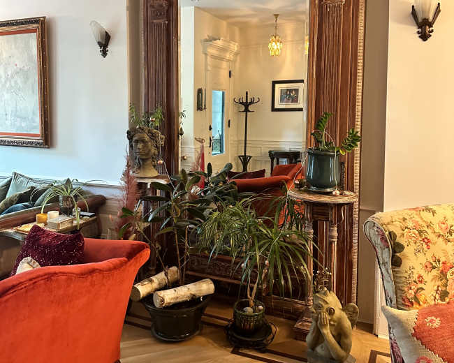 A large ornate mirror is framed by a decorative wooden border and surrounded by various plants and furniture in a cozy living room.