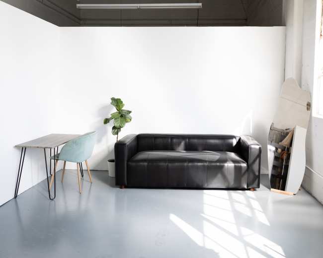 A black leather sofa is positioned against a white wall, accompanied by a small table, a light blue chair, a potted plant, and a surfboard leaning against the wall.