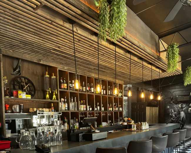 The image shows a modern bar with a black and white checkered floor, a long counter, and shelves stocked with various bottles, illuminated by hanging lights and surrounded by greenery.