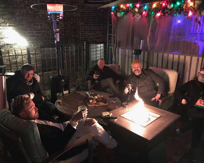 A group of five people sits around a lit fire pit on a patio, enjoying drinks and conversation under festive decorations.
