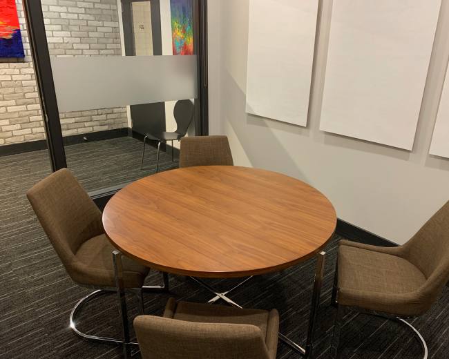 A round wooden table surrounded by four brown chairs in a modern office space with a brick wall and whiteboard surfaces.