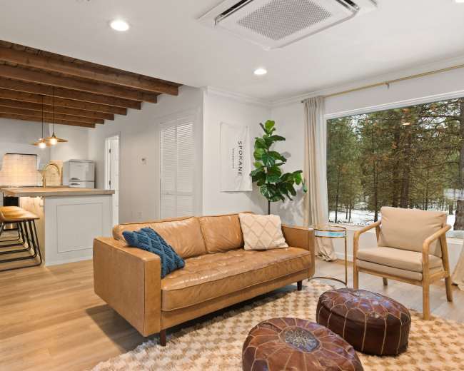 A cozy living space featuring a leather sofa, a wooden ceiling with exposed beams, and a kitchen area with bar seating next to a large window.