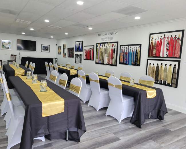 The image shows a room set up for an event with several long tables covered in black and gold tablecloths, surrounded by white chairs, and adorned with framed garments on the walls.