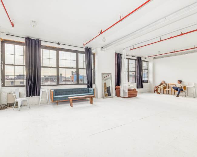 The image shows a spacious, empty industrial-style room with large windows, a few pieces of furniture, and exposed ceiling beams.