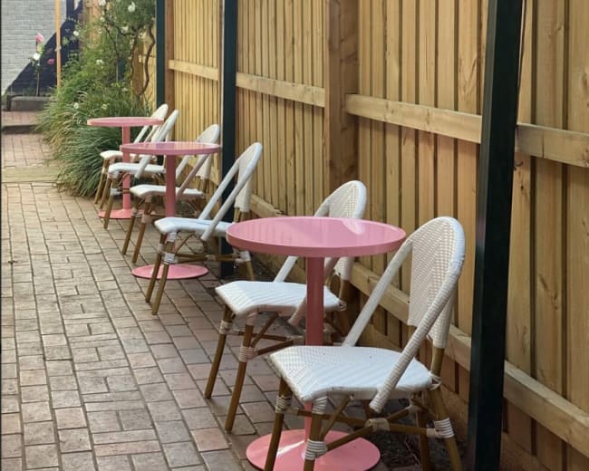 A row of white chairs with pink tables is placed against a wooden fence in an outdoor setting.