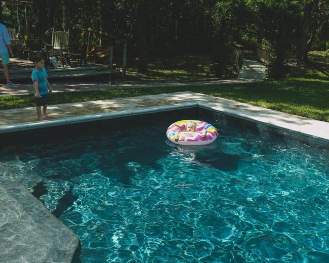 A child stands by the edge of a swimming pool with a colorful inflatable inner tube floating in the water.
