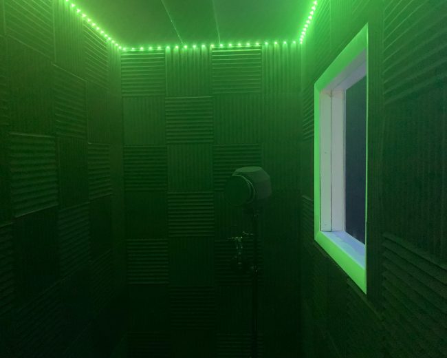 A small room with soundproof walls, illuminated by green LED lights, featuring a microphone stand and a window.