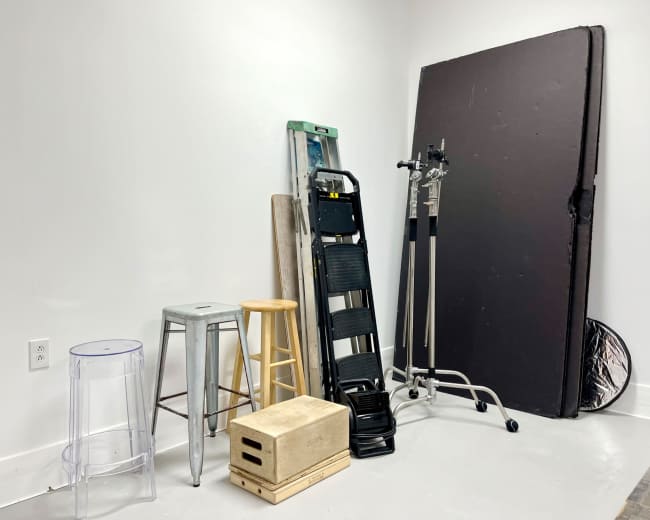 The image shows a collection of various items including stools, a ladder, and a large black board against a plain white wall.