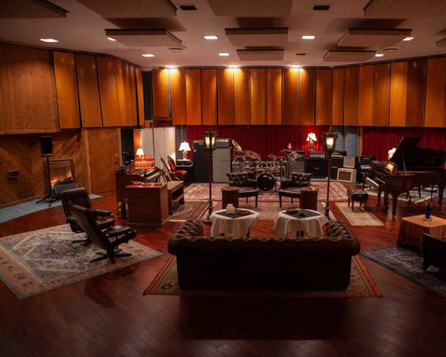 The image shows a spacious music recording studio with wooden paneling, vintage furniture, and various musical instruments arranged in the center.