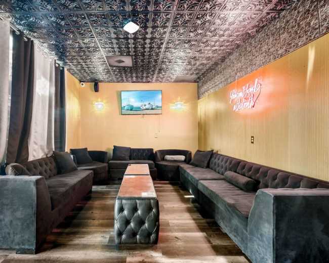 A spacious lounge area features dark velvet seating arranged around a central table, with a decorative ceiling and a wall-mounted television.